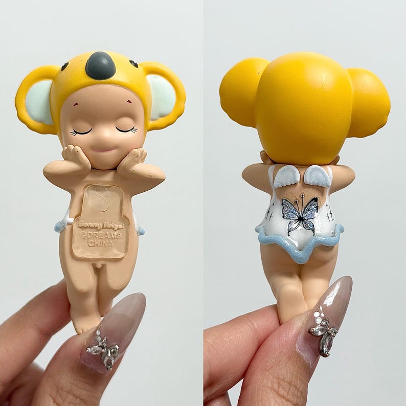 May include: A figurine of a child wearing a yellow koala hat and a light-coloured outfit. The figurine has closed eyes and is holding its hands up to its face. The back of the figurine has a butterfly design on the outfit.