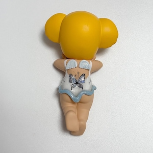 May include: A figurine of a person with yellow hair and a white and blue outfit. The back of the figurine is visible, showing a butterfly design on the outfit. The figurine is standing with its arms raised.