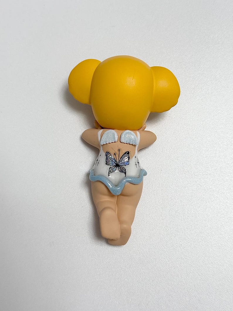 May include: A figurine of a person with yellow hair and a white and blue outfit. The back of the figurine is visible, showing a butterfly design on the outfit. The figurine is standing with its arms raised.