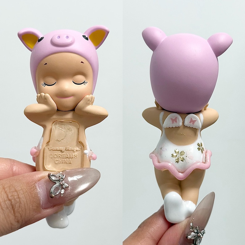 May include: A small figurine of a baby wearing a pink pig hat and a white swimsuit with a pink trim. The figurine is held up by a person's hand. The figurine has closed eyes and is smiling.