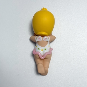 May include: A yellow duck-shaped figurine with a small tuft of feathers on its head. The figurine is wearing a white swimsuit with a pink trim and small daisy patterns. The figurine is facing away from the viewer.