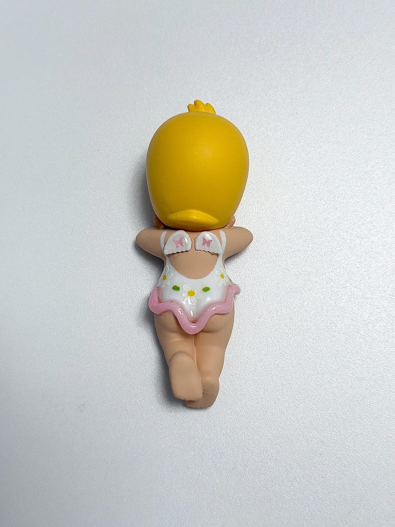 May include: A yellow duck-shaped figurine with a small tuft of feathers on its head. The figurine is wearing a white swimsuit with a pink trim and small daisy patterns. The figurine is facing away from the viewer.