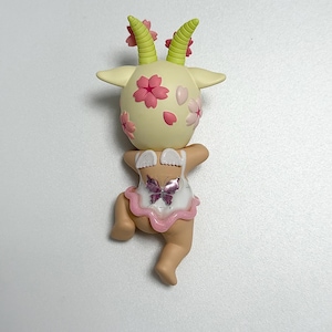 May include: A quirky figurine of a goat-like character with a cream-coloured head decorated with pink cherry blossoms and green horns. It wears a white outfit with a pink trim and a butterfly design. The figurine is in a playful pose.