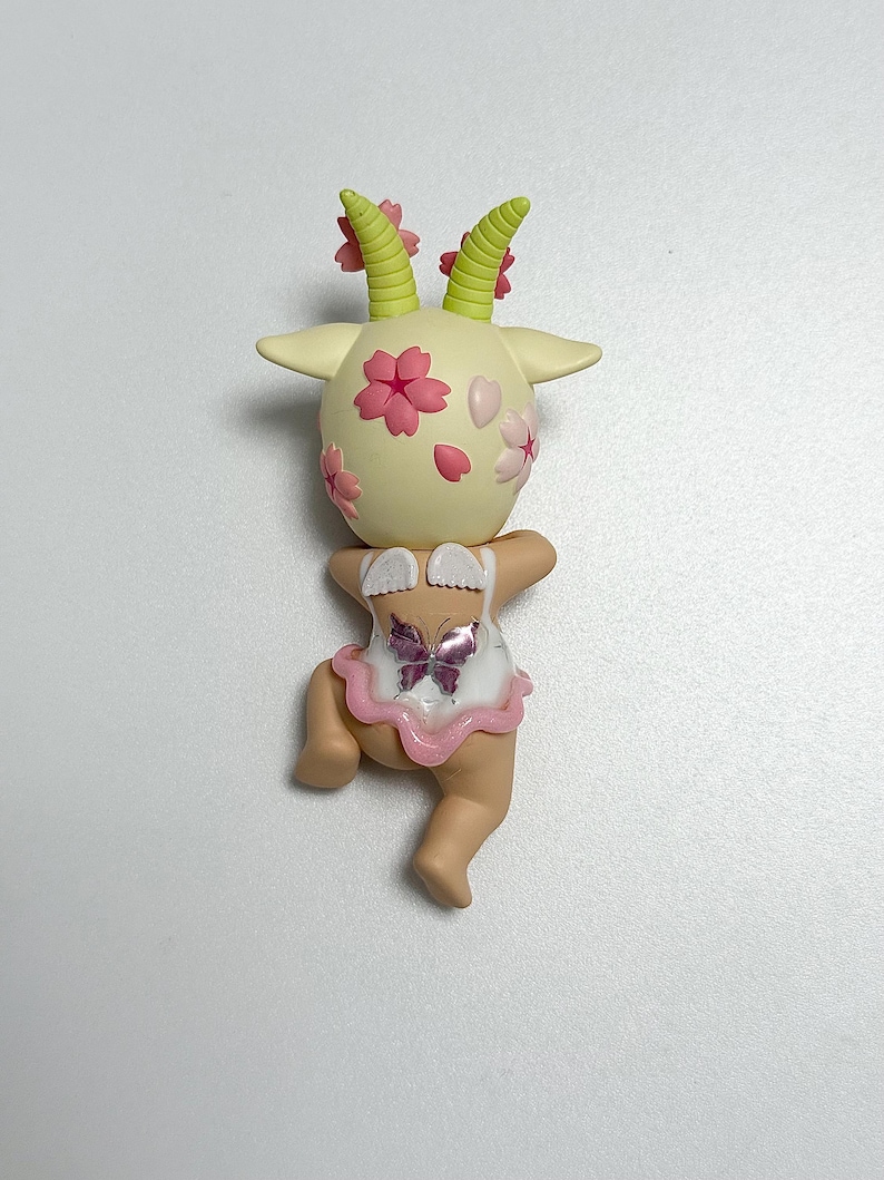 May include: A quirky figurine of a goat-like character with a cream-coloured head decorated with pink cherry blossoms and green horns. It wears a white outfit with a pink trim and a butterfly design. The figurine is in a playful pose.
