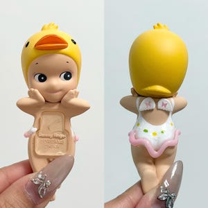 May include: A small figurine of a baby wearing a yellow duck hat and a white swimsuit with pink trim. The figurine has a smiling face and is holding its hands up to its cheeks. The text "SONNY ANGEL DREAMS CHINA" is visible on the figurine.