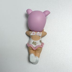 May include: A small figurine of a character with a pink head and ears, wearing a white swimsuit with a pink ruffle and floral accents. The figurine has a light skin tone and white socks.