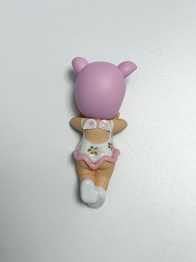 May include: A small figurine of a character with a pink head and ears, wearing a white swimsuit with a pink ruffle and floral accents. The figurine has a light skin tone and white socks.