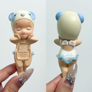 May include: A collectible figurine with a panda-style hat and a swimsuit with a cartoon character design. The figurine has closed eyes and is in a relaxed pose. The back of the figurine shows a white swimsuit with a blue trim and small wings. The text "DREAMS CHINA" is visible.
