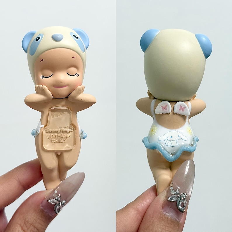 May include: A collectible figurine with a panda-style hat and a swimsuit with a cartoon character design. The figurine has closed eyes and is in a relaxed pose. The back of the figurine shows a white swimsuit with a blue trim and small wings. The text "DREAMS CHINA" is visible.