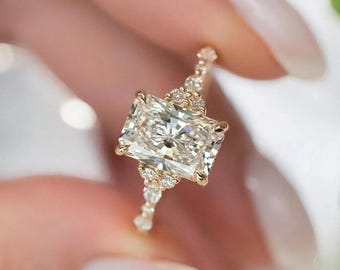 May include: A gold ring featuring a large, rectangular-cut diamond. The ring's band and setting are embellished with smaller diamonds. The ring is held between fingers, showcasing its intricate design and the brilliance of the central stone.