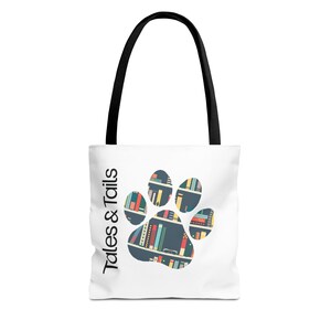 May include: White tote bag with black handles featuring a paw print design filled with colorful book illustrations. The text "Tales & Tails" is printed vertically on the side of the bag.
