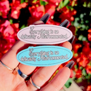 May include: Two oval stickers, one light pink and one light blue, with the text "Everything is so delicately interconnected" in a script font. The stickers are held up against a background of red flowers.