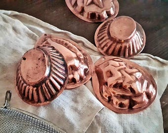 Puede incluir: Colección de moldes de cobre antiguos en varias formas, incluyendo una estrella, concha y diseños redondos. Los moldes se exhiben sobre un paño de color claro, mostrando su cálido tono marrón rojizo. Estos artículos de cocina vintage son ideales para hornear.