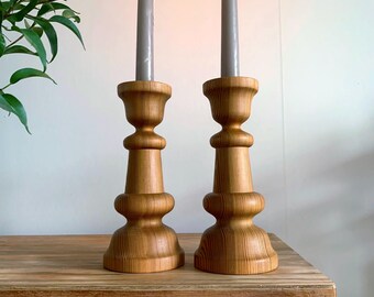 May include: A pair of wooden candle holders with a natural wood finish. The candle holders are tall and slender, with a flared base and a narrow top. The candle holders are made of solid wood and have a smooth, polished finish.