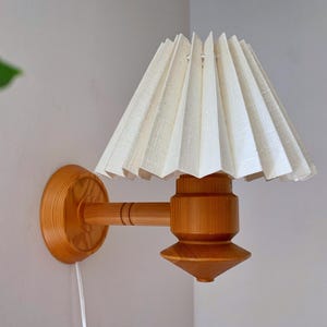 Może przedstawiać: Vintage lampa ścienna z plisowanym, kremowym abażurem. Lampa posiada drewniane ramię i podstawę, prezentując ciepły, naturalny odcień drewna. Design jest prosty i elegancki, odpowiedni do sypialni lub salonu.