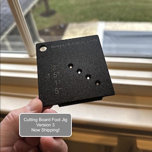 May include: Black cutting board foot jig with five holes and measurements in inches. The jig is labeled "Cutting Board Foot Jig Version 3 Now Shipping!" and "warpedboards.com".