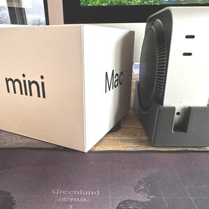 May include: A white box with the text "Mac mini" printed on it, next to a white Mac mini computer on a black stand.