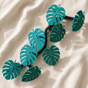 Op de afbeelding: Een decoratieve wandrankstructuur met een zwarte, gesegmenteerde stengel en zeven afzonderlijke, blauwgroene monstera-bladeren. De bladeren zijn verspringend langs de gebogen stengel geplaatst en vertonen gedetailleerde uitsparingen en nerven.