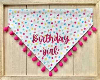 May include: A white dog bandana with a colorful confetti pattern and pink pom-poms around the edges. The bandana has the text "Birthday girl" in pink lettering.