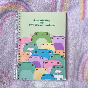 May include: A green spiral bound notebook with the text "Just minding my own sticker business" and a group of colorful frogs on the cover.