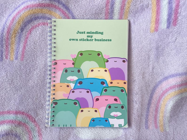 May include: A green spiral bound notebook with the text "Just minding my own sticker business" and a group of colorful frogs on the cover.