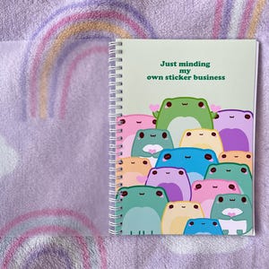 May include: A green spiral bound notebook with a light green background and the text "Just minding my own sticker business". The cover features a group of cartoon frogs in various colors and poses.