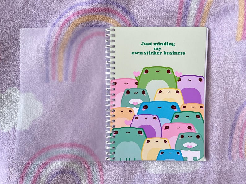 May include: A green spiral bound notebook with a light green background and the text "Just minding my own sticker business". The cover features a group of cartoon frogs in various colors and poses.
