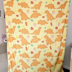 Könnte beinhalten: Eine gelbe Decke mit einem sich wiederholenden Muster aus orangefarbenen Zeichentrick-Dinosauriern, Cupcakes, Ketchupflaschen und anderen Lebensmitteln. Die Decke besteht aus einem weichen Material und eignet sich für die Verwendung auf einem Bett oder Sofa.