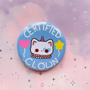 Könnte beinhalten: Ein blauer und weißer Knopf mit dem Text "CERTIFIED CLOWN" und einer Comic-Katze, die einen Partyhut trägt und einen Herz- und Sternballon hält.