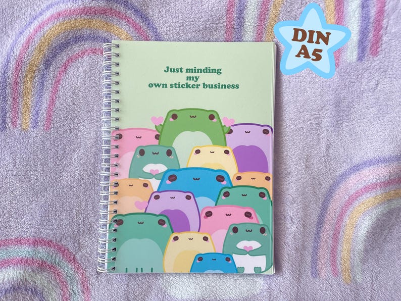 May include: A green spiral bound notebook with the text "Just minding my own sticker business" on the front cover. The cover features a colorful illustration of frogs with hearts.