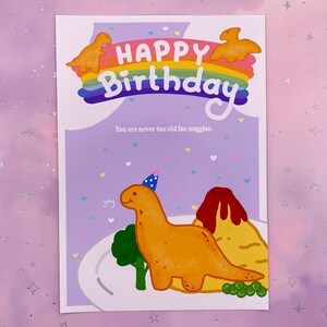 以下が含まれることがあります： 虹と「Happy Birthday」の文字が書かれた紫色のカード。カードには、パーティーハットをかぶった恐竜の漫画と、火山型のナゲットも描かれている。「You are never too old for nuggets.」という文字がカードに印刷されている。