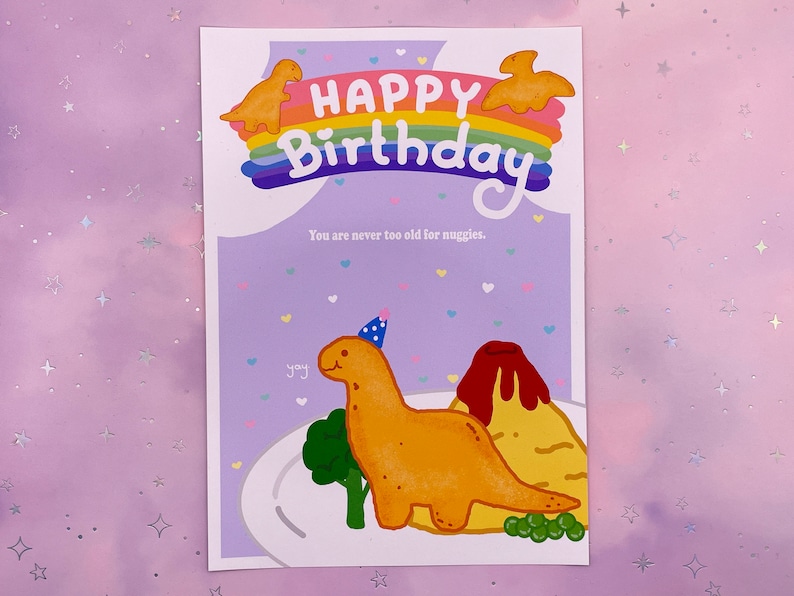 以下が含まれることがあります： 虹と「Happy Birthday」の文字が書かれた紫色のカード。カードには、パーティーハットをかぶった恐竜の漫画と、火山型のナゲットも描かれている。「You are never too old for nuggets.」という文字がカードに印刷されている。