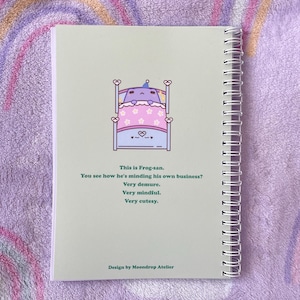 May include: A green and white notebook with a spiral binding. The cover features a cartoon frog in bed with the text "This is Frog-san. You see how he's minding his own business? Very demure. Very mindful. Very cutesy." and "Design by Moondrop Atelier".