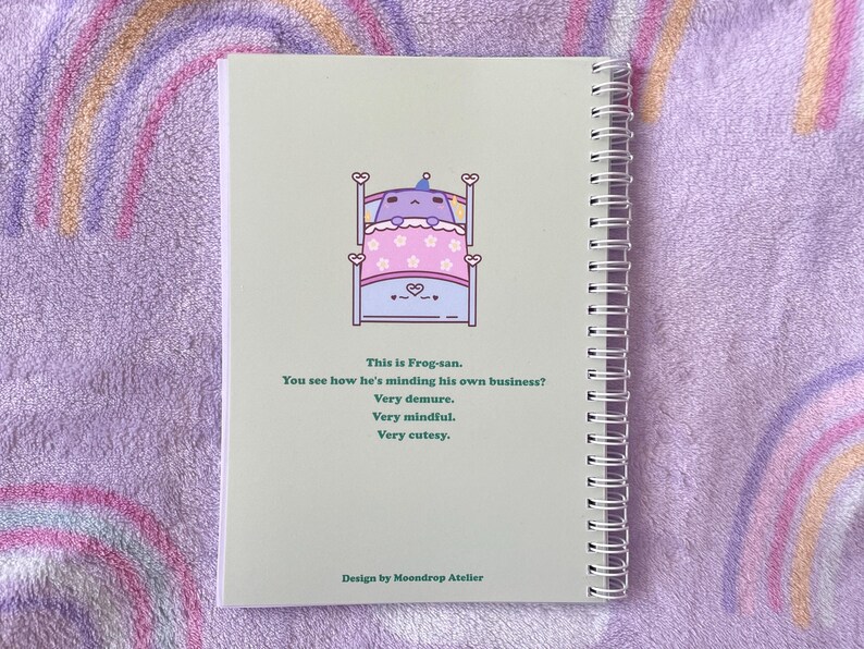 May include: A green and white notebook with a spiral binding. The cover features a cartoon frog in bed with the text "This is Frog-san. You see how he's minding his own business? Very demure. Very mindful. Very cutesy." and "Design by Moondrop Atelier".