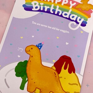 以下が含まれることがあります： 虹と「Happy Birthday」の文字が書かれた誕生日カード。カードには、パーティーハットをかぶって料理の入った皿を持っている漫画の恐竜が描かれている。「You are never too old for nuggets.」という文字がカードに印刷されている。