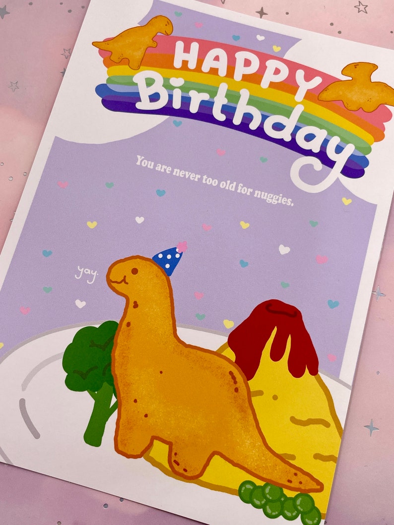 以下が含まれることがあります： 虹と「Happy Birthday」の文字が書かれた誕生日カード。カードには、パーティーハットをかぶって料理の入った皿を持っている漫画の恐竜が描かれている。「You are never too old for nuggets.」という文字がカードに印刷されている。