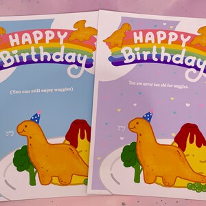 以下が含まれることがあります： パーティーハットをかぶった恐竜と虹が描かれた、カラフルな誕生日カード2枚。カードには「ハッピーバースデー」の文字と、「You can still enjoy nuggies」と「You are never too old for nuggies.」のフレーズが書かれています。