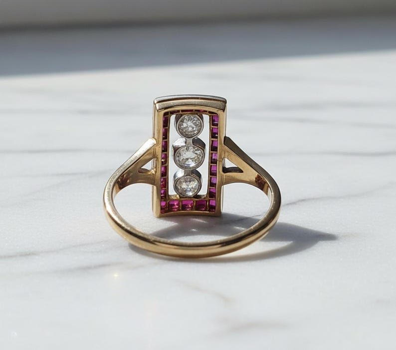 Pu&ograve; includere: Un anello in oro con una testa rettangolare caratterizzata da una fila verticale di tre diamanti rotondi taglio brillante incastonati in metallo bianco. La cornice rettangolare &egrave; bordata da piccoli rubini rossi taglio quadrato su una fascia in oro giallo lucido.