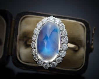 May include: An oval-shaped ring with a large, iridescent blue gemstone, surrounded by a halo of small, clear diamonds. The ring is set in a gold band and presented in a vintage jewelry box.