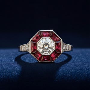 Puede incluir: Un anillo de estilo vintage con una gran piedra central transparente de talla redonda. El anillo presenta un diseño geométrico con un halo de piedras rojas de talla cuadrada. La banda es plateada y tiene pequeñas piedras de acento.