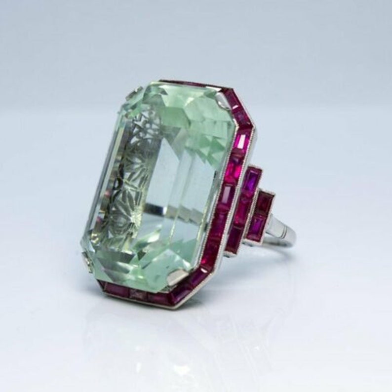 May include: A vintage ring featuring a large, light green emerald-cut aquamarine stone surrounded by a border of small, rectangular ruby stones set in platinum.