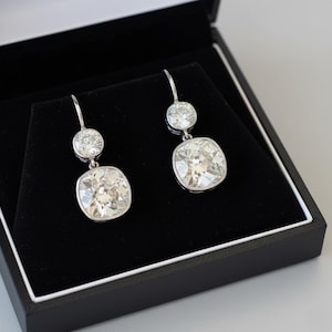 May include: A pair of silver-toned drop earrings, each featuring two cushion-cut clear stones set in metal. The earrings are displayed inside a black jewelry box with a white interior lining, showcasing a reflective, faceted design.