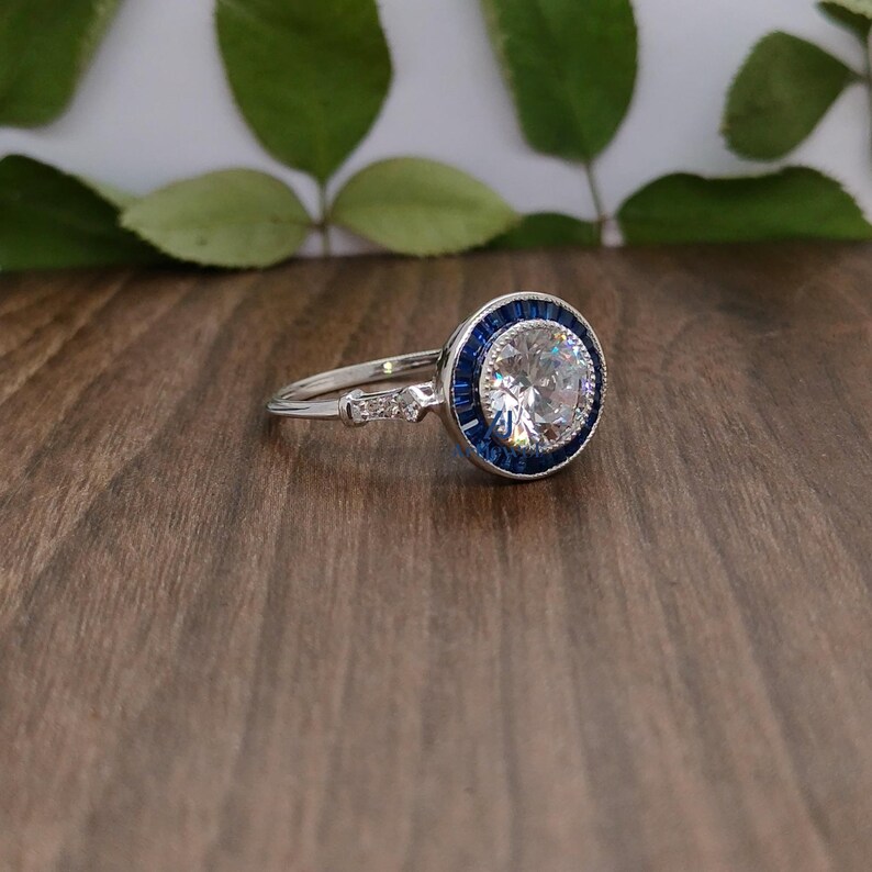 May include: A silver ring featuring a large, clear, round central stone encircled by a halo of small, blue gemstones. The band has additional small stones. The ring is set against a wooden surface with green leaves in the background.