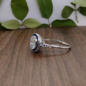 May include: A silver ring featuring a large, clear, round central stone. The ring has a blue border around the main stone and smaller stones along the band. It rests on a wooden surface, with green leaves in the background.