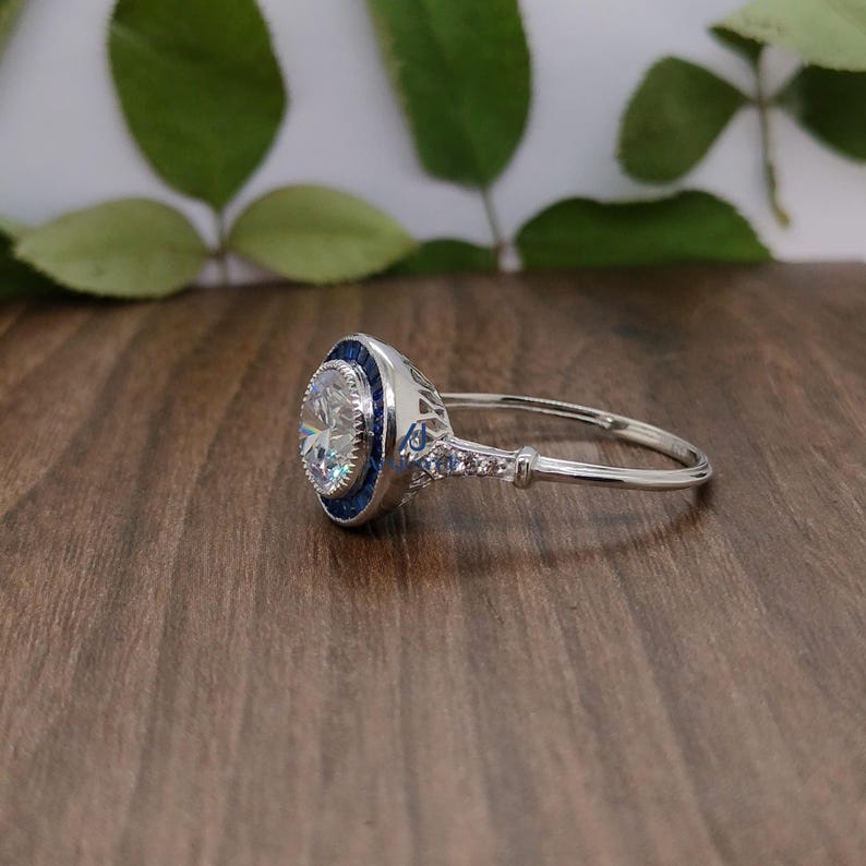 May include: A silver ring featuring a large, clear, round central stone. The ring has a blue border around the main stone and smaller stones along the band. It rests on a wooden surface, with green leaves in the background.