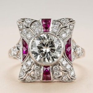 Peut inclure: Une bague de style vintage avec une grosse pierre centrale en diamant clair et rond. La bague présente de petits diamants et des accents de rubis rectangulaires sertis dans un sertissage en métal argenté orné. Le design est symétrique et détaillé.