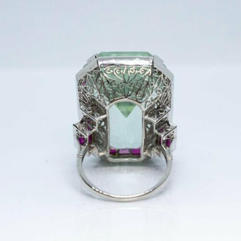 May include: A vintage platinum ring featuring a large, emerald-cut aquamarine stone surrounded by a delicate filigree design. The ring is accented with small ruby stones.