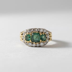 May include: A gold ring with three square emeralds at its centre, encircled by small diamonds. The ring has a classic design with a yellow gold band. The background is white.