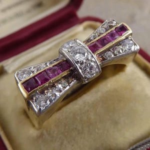 May include: A bow-shaped ring with a gold band, featuring a central section of square-cut rubies and surrounding diamonds. The ring is set in a silver and gold setting, showcasing a vintage design.