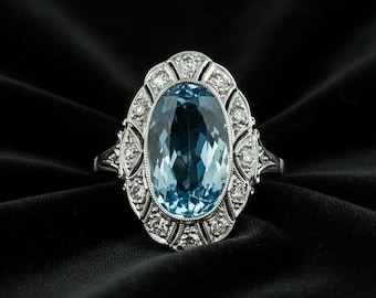May include: An oval-shaped, light blue gemstone ring with a silver band. The gemstone is surrounded by smaller, clear stones and ornate silver detailing. The ring is set against a black background, highlighting its elegant design.